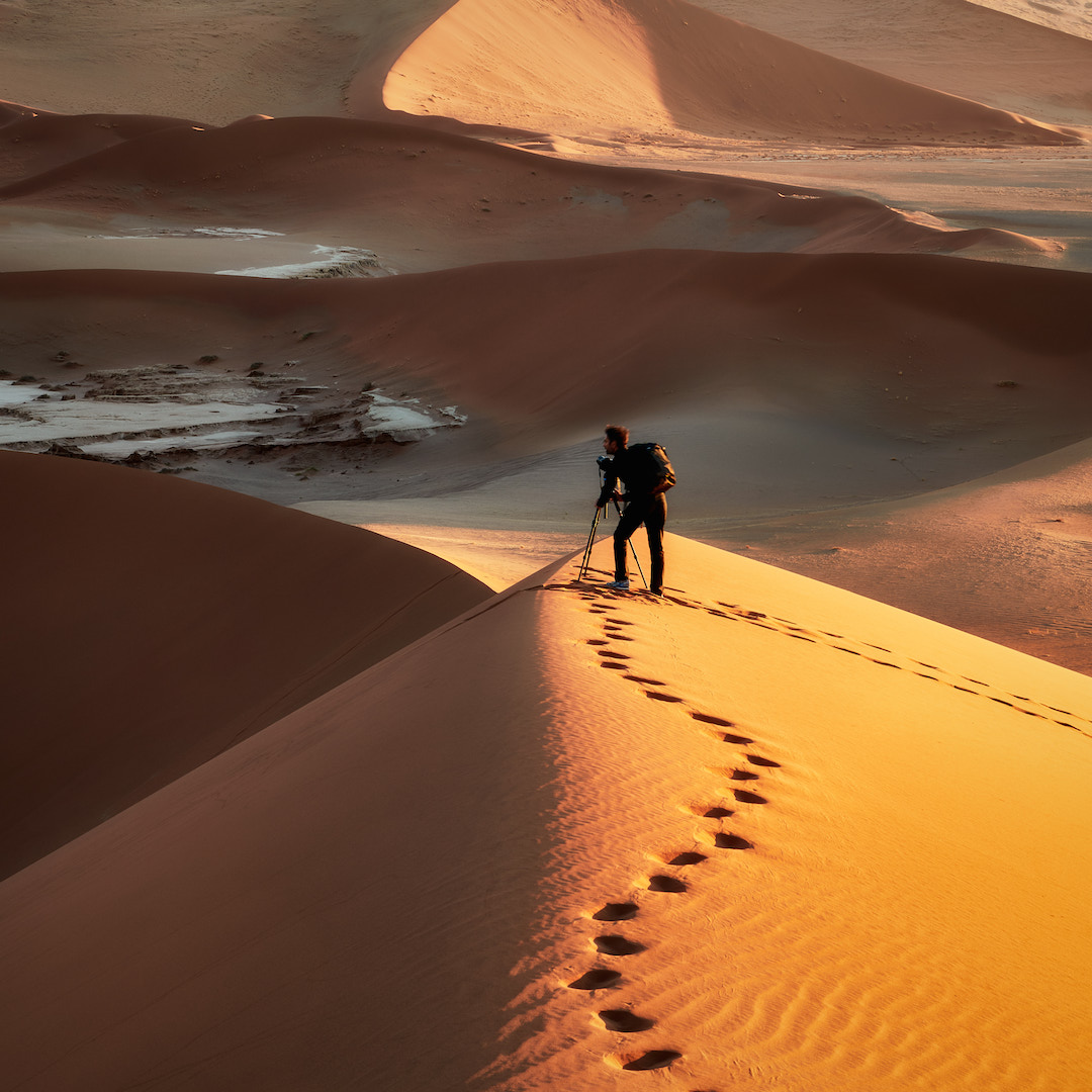 photographer stands on top of a sand dune, capturing the desert landscape, with footprints trailing behind them