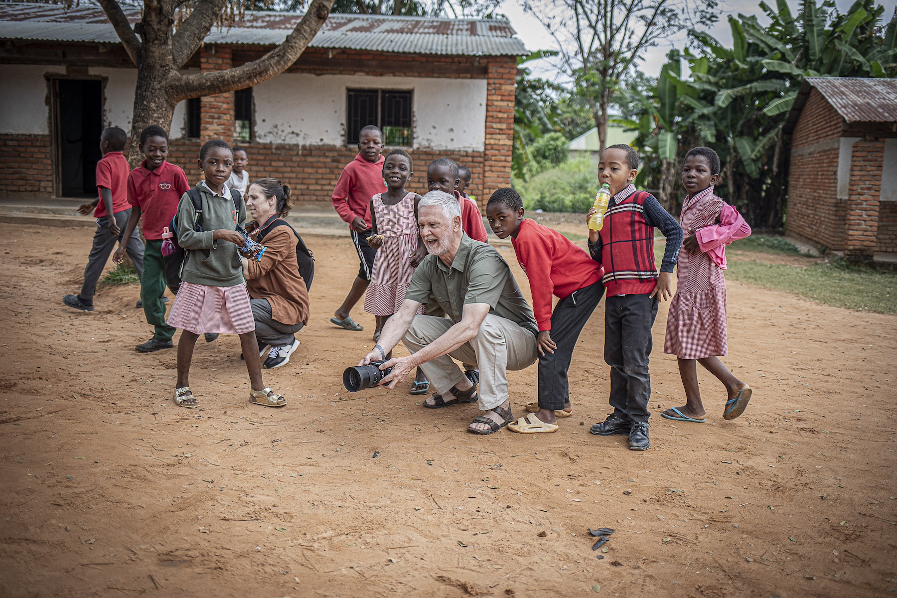 A crouching adult with a camera, surrounded by children and another adult in a village.
