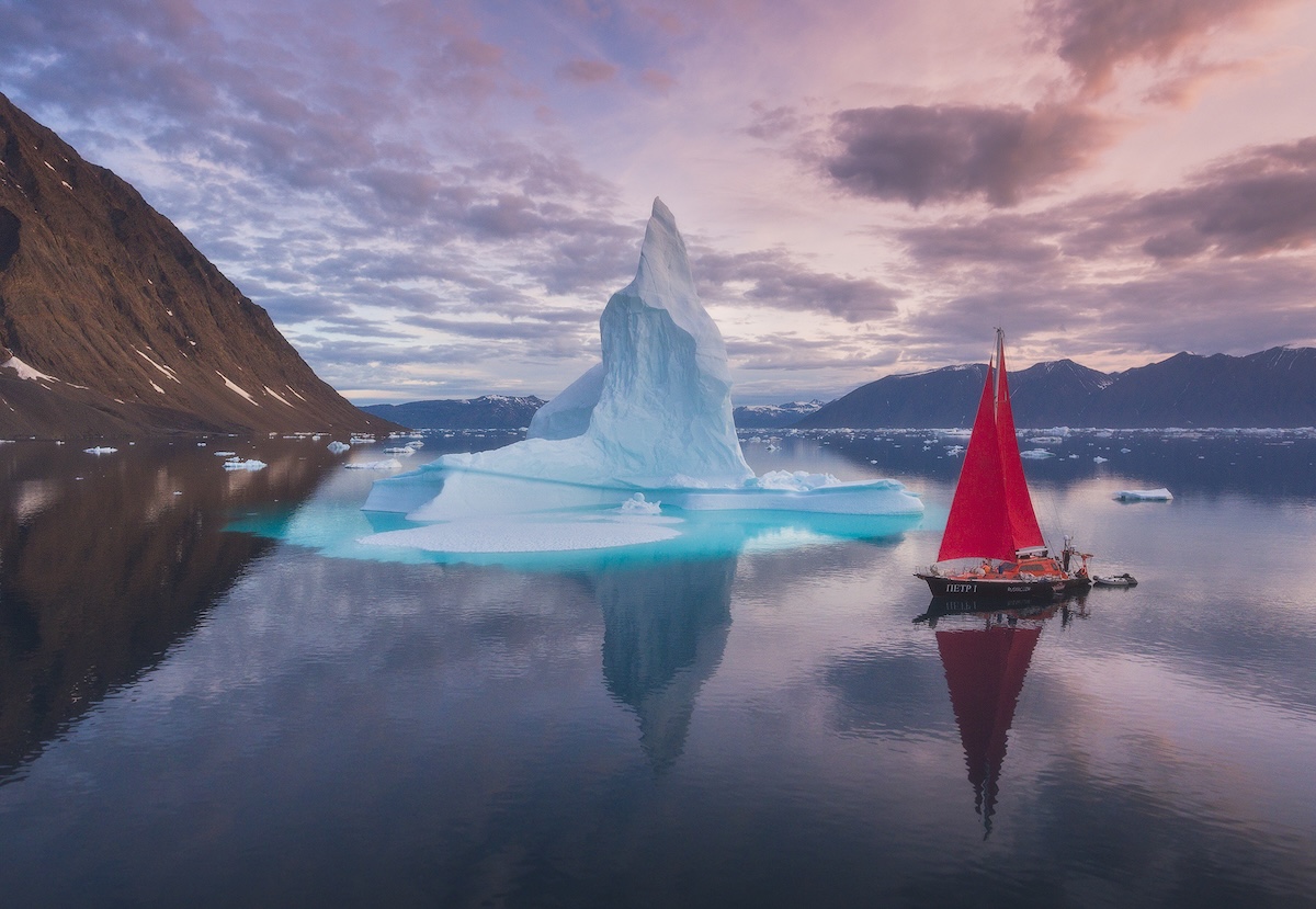 Sailboat with red sails near a towering iceberg in calm Arctic waters