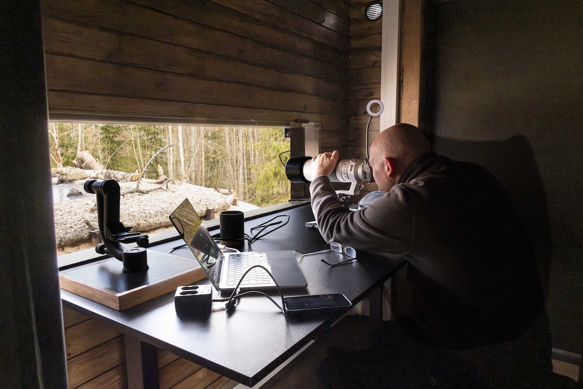 A person using a large camera lens to photograph wildlife from inside a wooden hide