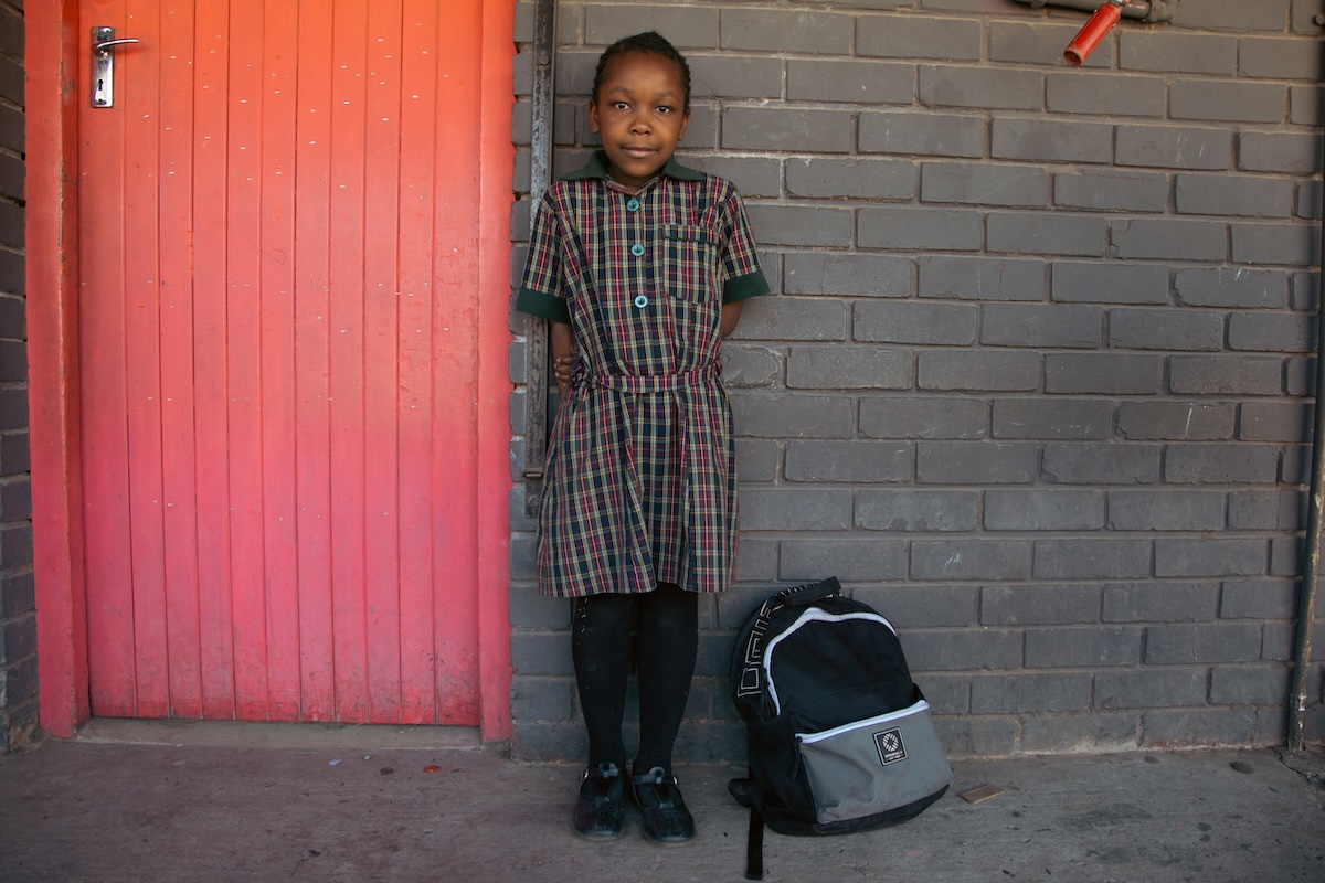 Young girl in a plaid dress standing by a wall with her backpack