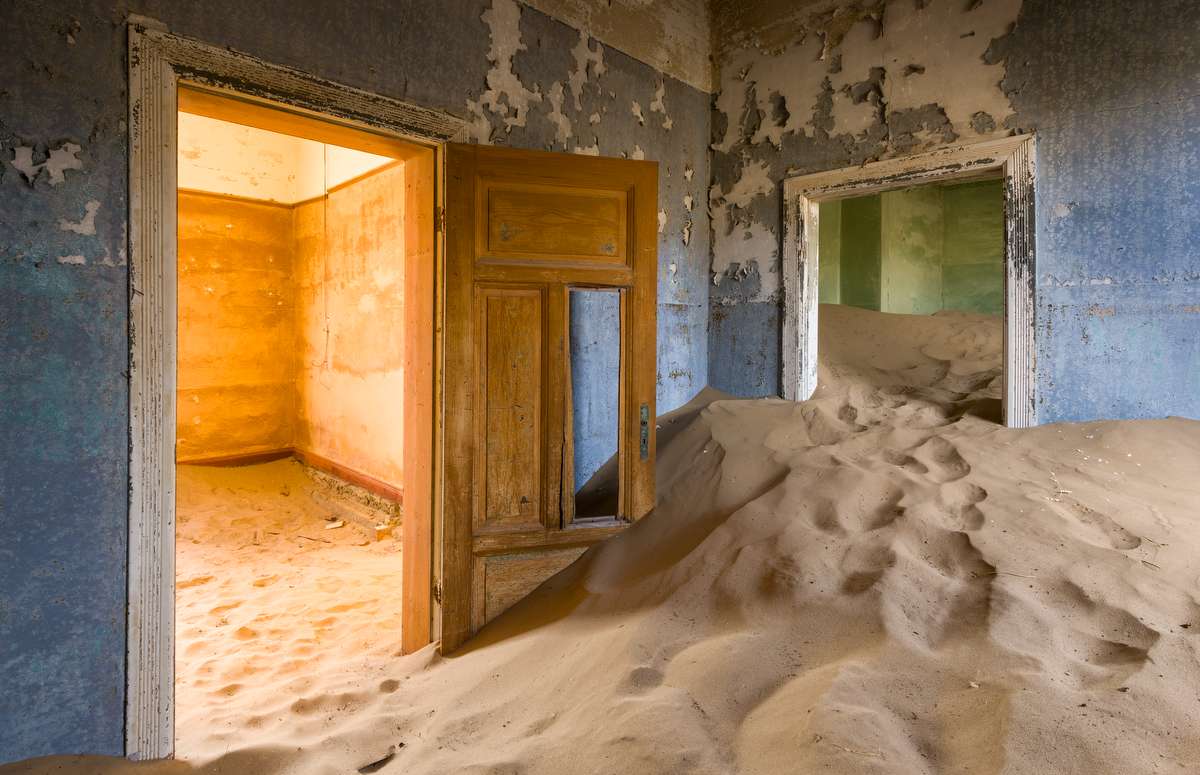 Sand drifts through rooms with peeling blue paint and broken doors in an abandoned building