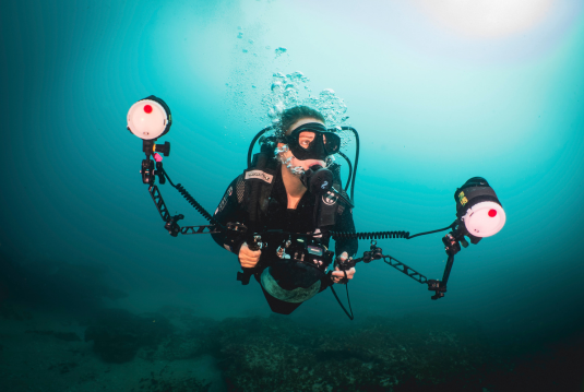 A diver equipped with a camera rig and dual strobe lights ascends through sunlit blue water, showcasing professional underwater photography gear used during an Underwater Photography Workshop on a Penda Photo Tour.