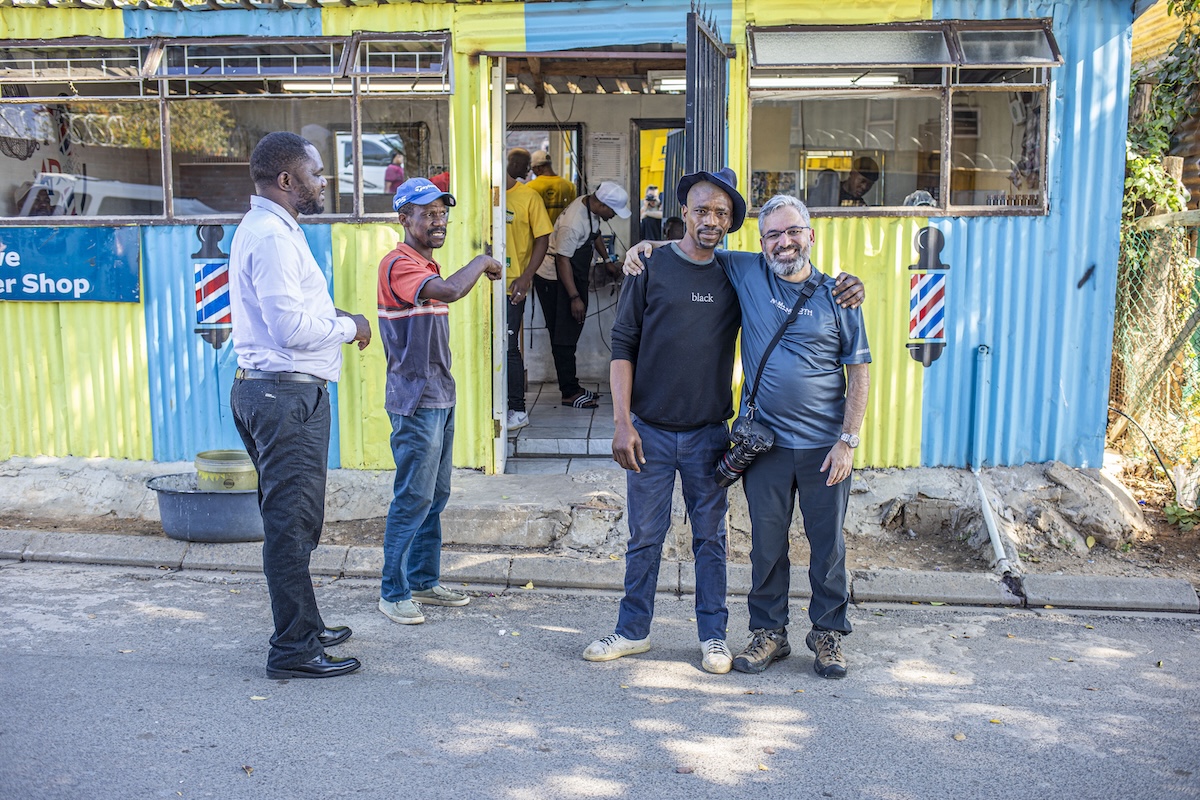 Two men smiling in front of a bright barbershop, with others nearby