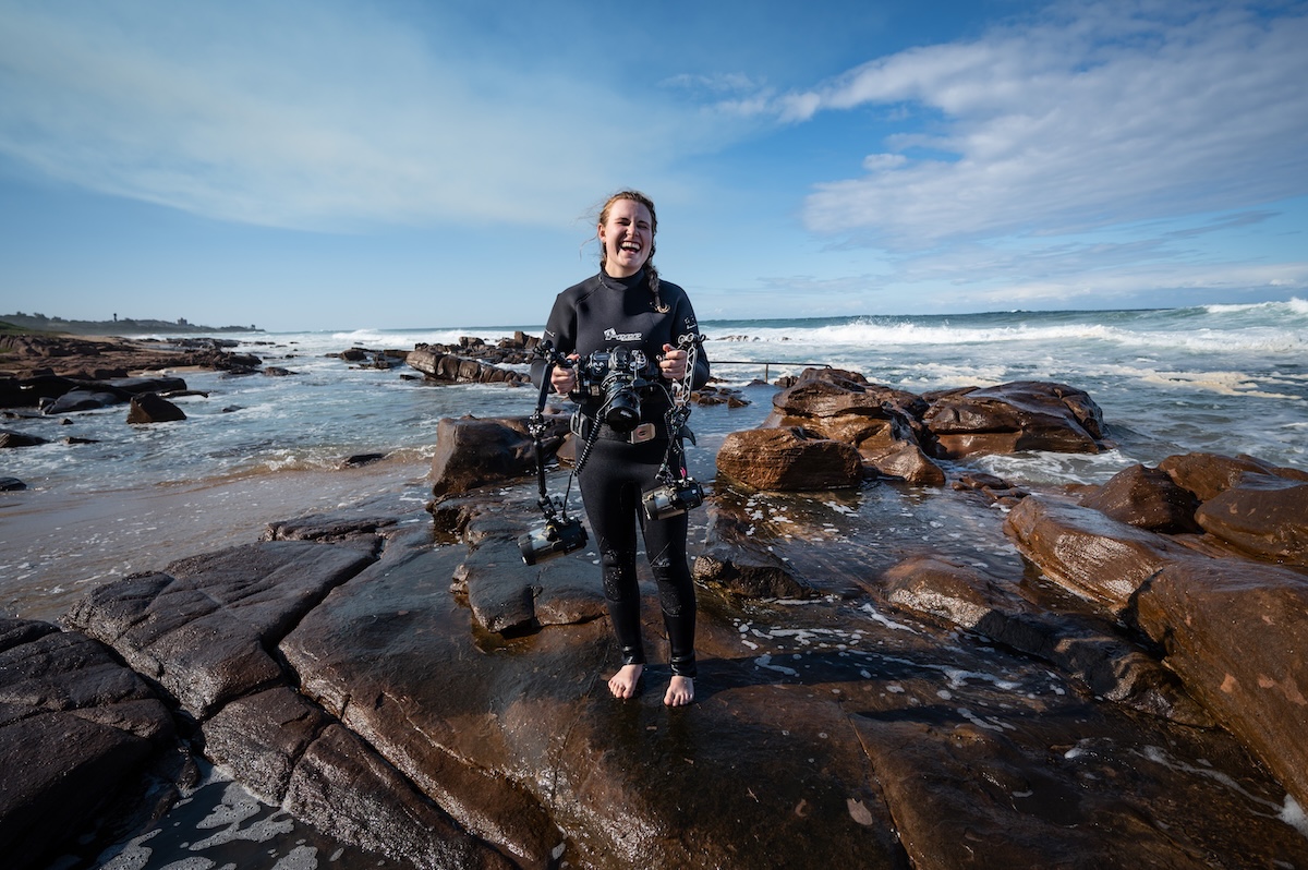 Woman in wetsuit holding underwater camera on rocky shore