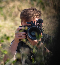 A photographer crouched low with a telephoto lens, framing a shot through tall grass, captured during a Wildlife Photography Internship on a Penda Photo Tour.