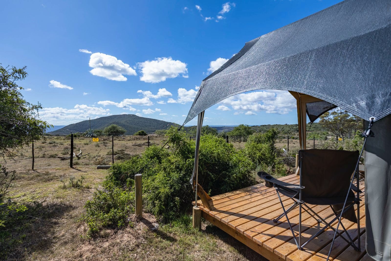 Outdoor camping setup with a canvas tent on a wooden platform, a folding chair in front, surrounded by dry grass and a wire fence, with a mountain in the background under a bright blue sky with scattered clouds.