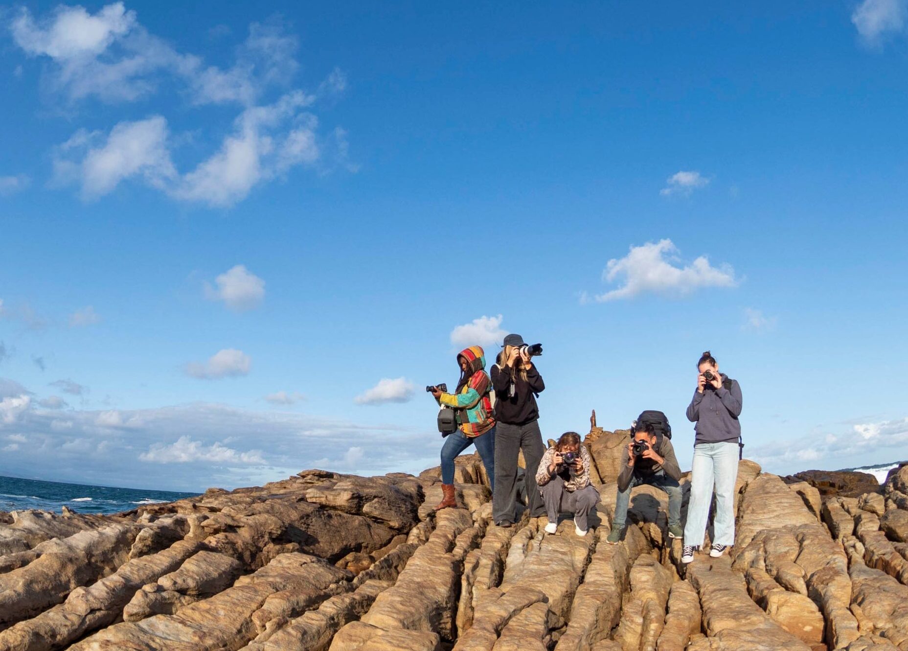 Five photographers on a rocky coastal outcrop, holding cameras and capturing the ocean view under a bright blue sky with scattered clouds.