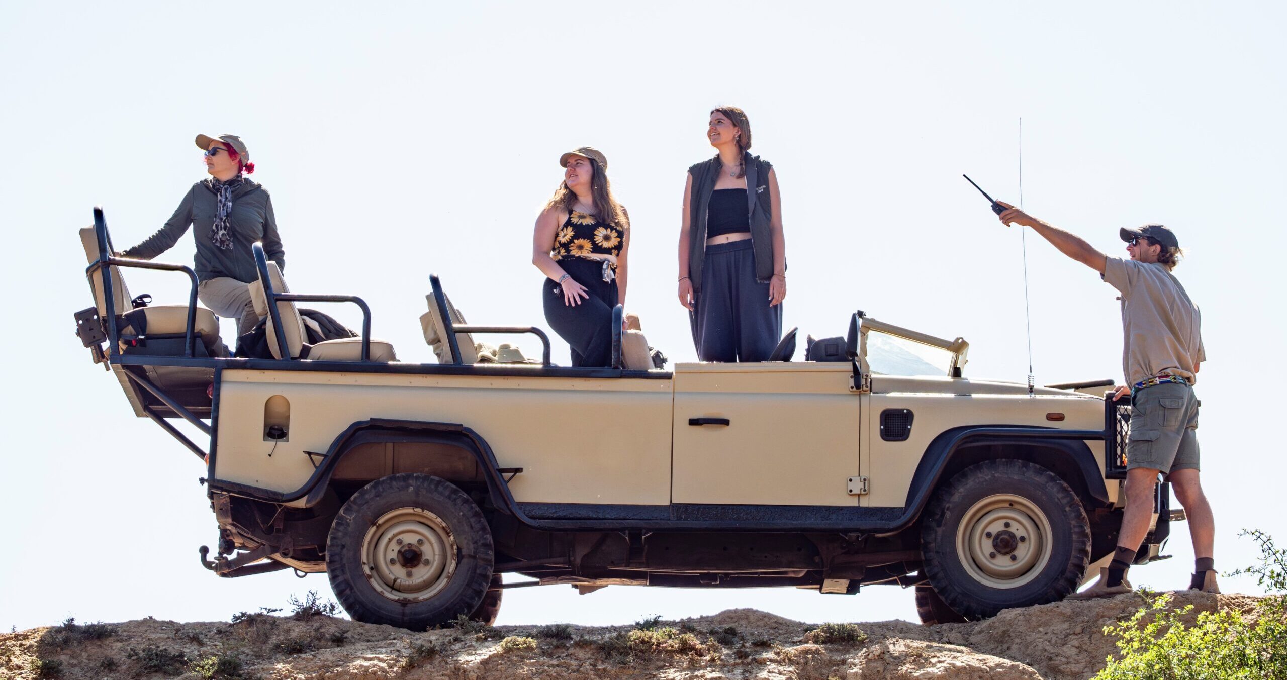 Safari-style open-top vehicle parked on rocky terrain, with three people observing from inside and one guide standing outside pointing into the distance under a bright sky.
