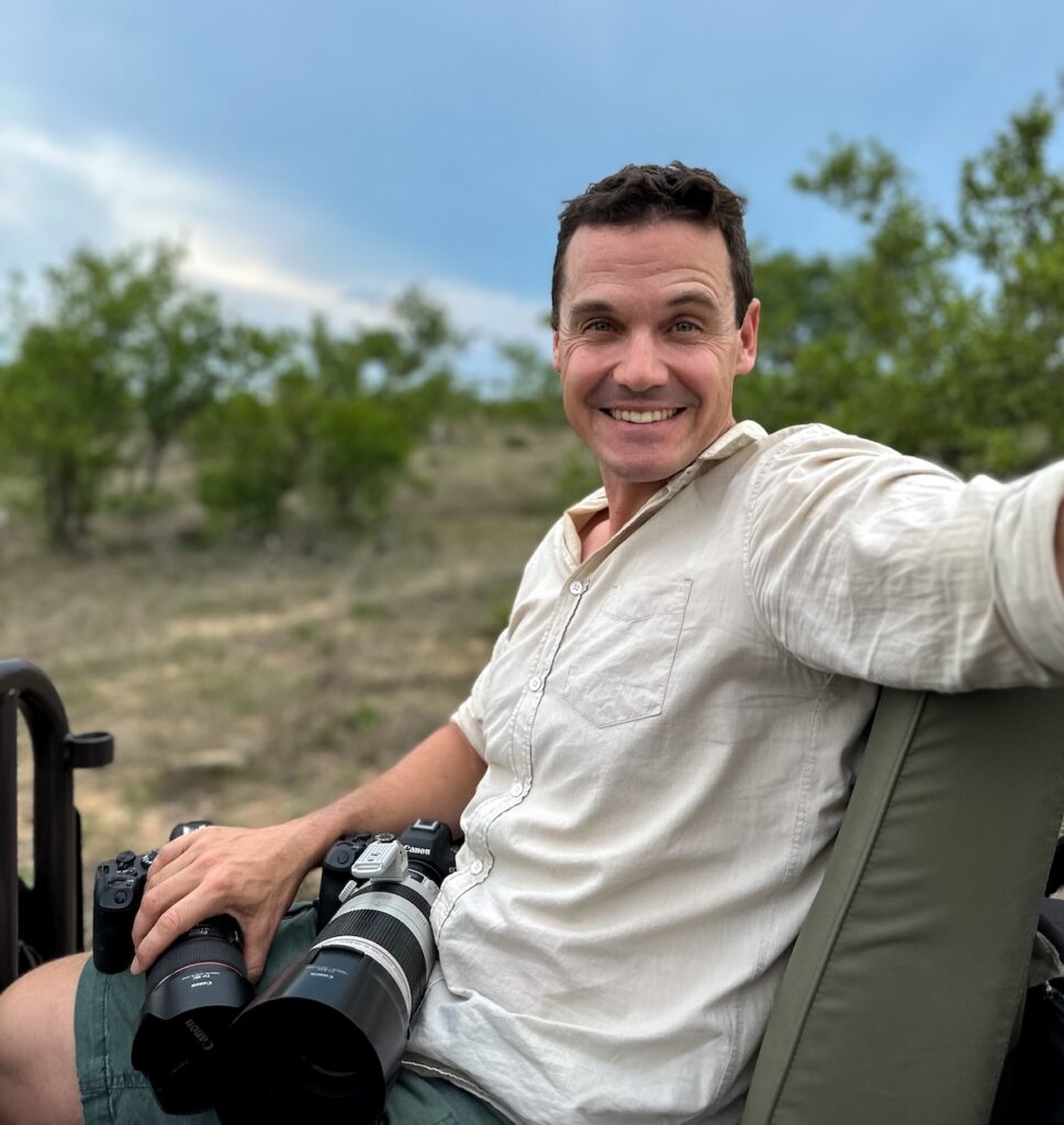 Penda Photo Tours guide Mike Dexter smiles from a safari vehicle seat, holding two Canon cameras with telephoto lenses against a green bushveld backdrop.