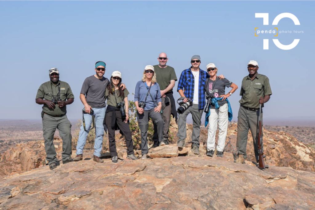 A group of guides and photographers pose together with cameras and binoculars atop a rocky outcrop overlooking the bush, marking Penda Photo Tours' 10th anniversary.